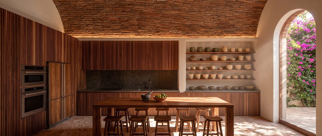 Visualización de cocina bajo bóveda de ladrillo con gabinetes de madera acanalada, mesa larga de madera y repisas con cerámica